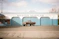 A vintage pickup truck is parked in front of a blue and white automotive shop called Master Muffler. The building has large, closed garage doors and signage that includes services like catalytic converters, brakes, shocks, and struts. The sky is partly cloudy, and there are no people present.