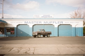A vintage pickup truck is parked in front of a blue and white automotive shop called Master Muffler. The building has large, closed garage doors and signage that includes services like catalytic converters, brakes, shocks, and struts. The sky is partly cloudy, and there are no people present.