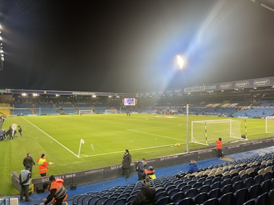 A well-lit football stadium with green grass and seating areas. Several people are present around the field, some wearing high-visibility jackets. The stands have numerous empty blue seats and screens display information. Stadium lighting creates a bright atmosphere.