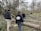 A group of individuals are engaged in clearing fallen trees and branches in a wooded area. They appear to be part of a work crew, wearing casual outdoor clothing and gloves. The surroundings are dense with leafless trees, and the ground is scattered with tree debris, indicating recent storm damage.