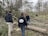 A group of individuals are engaged in clearing fallen trees and branches in a wooded area. They appear to be part of a work crew, wearing casual outdoor clothing and gloves. The surroundings are dense with leafless trees, and the ground is scattered with tree debris, indicating recent storm damage.