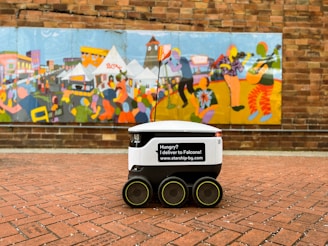 A delivery robot with six wheels is positioned on a brick-paved sidewalk in front of a colorful mural. The mural depicts various stylized figures and scenes in vibrant colors, resembling an outdoor festival or market with tents and musicians. The robot has a flag and a message promoting deliveries.