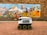 A delivery robot with six wheels is positioned on a brick-paved sidewalk in front of a colorful mural. The mural depicts various stylized figures and scenes in vibrant colors, resembling an outdoor festival or market with tents and musicians. The robot has a flag and a message promoting deliveries.