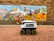 A delivery robot with six wheels is positioned on a brick-paved sidewalk in front of a colorful mural. The mural depicts various stylized figures and scenes in vibrant colors, resembling an outdoor festival or market with tents and musicians. The robot has a flag and a message promoting deliveries.