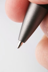 A close-up view of the tip of a metallic pen held between two fingers, with visible fingerprint details.