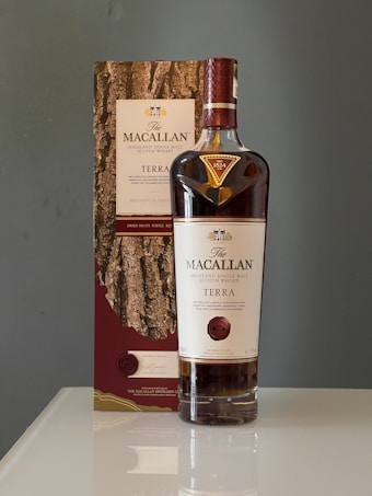 A bottle of The Macallan Terra Highland Single Malt Scotch Whisky is placed next to its packaging box, which features a wood texture design. The bottle has a dark brown and red label with gold accents. Both the bottle and the box are on a glossy surface with a neutral background.