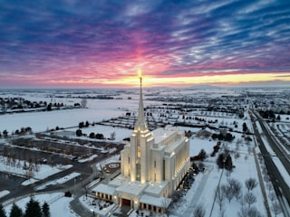 Aerial view of a large, illuminated temple with a tall spire, surrounded by snow-covered ground and a sprawling suburban area. The sun is setting, casting a purple and pink hue across the clouds in the sky.