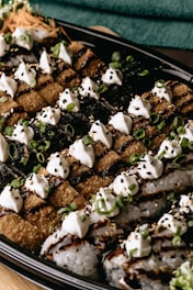 An appetizing presentation of sushi rolls adorned with creamy dollops and sprinkled with sesame seeds and chopped green onions. The sushi is arranged neatly on a black platter, with a mixture of crispy and soft textures, drizzled with a dark sauce.