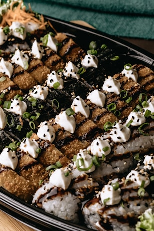 An appetizing presentation of sushi rolls adorned with creamy dollops and sprinkled with sesame seeds and chopped green onions. The sushi is arranged neatly on a black platter, with a mixture of crispy and soft textures, drizzled with a dark sauce.
