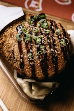 A deliciously golden-brown deep-fried dish garnished with green onions and sesame seeds. Drizzled with a dark sauce, it sits atop some white pickled vegetables in a rectangular dish on a wooden surface.