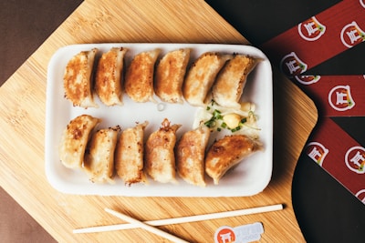 A rectangular white plate with twelve pan-fried dumplings arranged neatly. The dumplings are golden-brown, and there is a small garnish of diced green onions and a dollop of mayonnaise beside them. Two wooden chopsticks are placed on a wooden cutting board next to the plate. Red paper strips with Asian-themed graphics are scattered around.