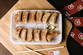 A rectangular white foam tray holds perfectly arranged pan-fried dumplings, golden-brown in color, served with a side of chopped green onions and a dollop of sauce. The tray is placed on a wooden board, accompanied by what looks like a pair of chopsticks and a partial view of a branded napkin.