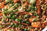 A close-up of a dish featuring stir-fried noodles mixed with vegetables such as carrots and green onions, as well as pieces of shrimp. The noodles are coated in a savory sauce and garnished with freshly chopped green onions.