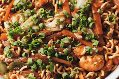 A close-up of a dish featuring stir-fried noodles mixed with vegetables such as carrots and green onions, as well as pieces of shrimp. The noodles are coated in a savory sauce and garnished with freshly chopped green onions.