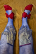 A pair of legs wearing blue fishnet stockings with red bow shoes, with two gardening gloves placed over the knees. The legs are resting on a mustard yellow fabric background.
