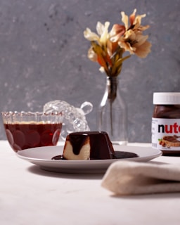 A small dessert covered in chocolate sauce, placed on a white plate, is positioned at the forefront on a light-colored surface. In the background, there is a jar of Nutella partially visible, a glass container filled with a dark liquid, and a clear glass bottle with a few dried flowers.