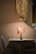 A cozy restaurant setting featuring a small table with a decorative vase of vibrant flowers placed at its center. The table is set for two, with utensils neatly arranged. Above the table hangs a modern brass pendant light, casting a warm glow on the scene. The background includes textured walls and a hint of floral decor.