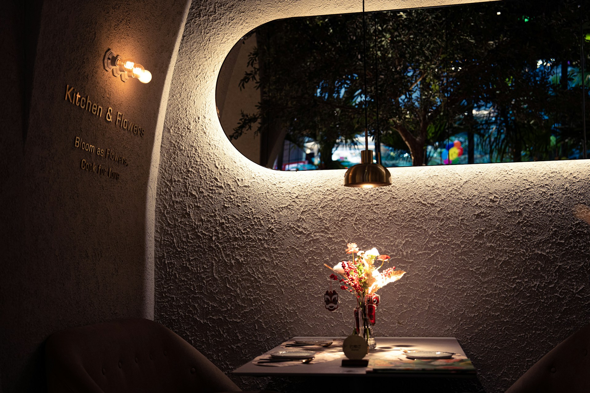 A cozy dining setting featuring a textured wall with 'Kitchen & Flowers' signage and a motivational phrase. A round window provides a view of foliage outside. A small table with two elegant chairs is centrally placed, adorned with a colorful flower arrangement. The scene is illuminated by a warm light bulb fixture and a stylish pendant light above the table.