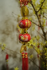 An ornamental decor with red and gold colors hangs against a backdrop of branches adorned with yellow flowers. The ornament features intricate patterns and tassels with Chinese characters.