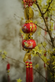An ornamental decor with red and gold colors hangs against a backdrop of branches adorned with yellow flowers. The ornament features intricate patterns and tassels with Chinese characters.