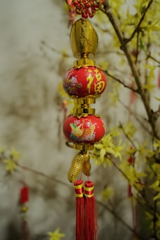 An ornamental decor with red and gold colors hangs against a backdrop of branches adorned with yellow flowers. The ornament features intricate patterns and tassels with Chinese characters.
