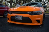 A bright orange sports car, likely a Dodge Charger SRT, is parked on a shadowed asphalt surface. The car features a black front grille and a license plate that reads 'KILLER CAT' with Florida written on it. A red car is partially visible on the left side, and trees can be seen in the background with a person standing nearby.