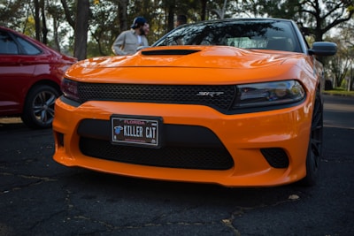 A bright orange sports car, likely a Dodge Charger SRT, is parked on a shadowed asphalt surface. The car features a black front grille and a license plate that reads 'KILLER CAT' with Florida written on it. A red car is partially visible on the left side, and trees can be seen in the background with a person standing nearby.
