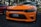A bright orange sports car, likely a Dodge Charger SRT, is parked on a shadowed asphalt surface. The car features a black front grille and a license plate that reads 'KILLER CAT' with Florida written on it. A red car is partially visible on the left side, and trees can be seen in the background with a person standing nearby.