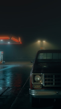 A dimly lit gas station at night with wet pavement reflecting the lights. A vintage red truck is parked on the right side, partially cut off from the frame. The atmosphere is foggy and eerie, with a sense of isolation.