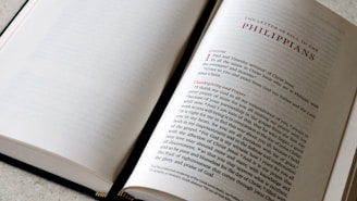 An open book lying on a light-colored surface, with the left page mostly blank while the right page contains printed text. The heading 'The Letter of Paul to the Philippians' is prominently displayed at the top of the right page in capital letters. The text on the page appears to be a passage from the Bible, beginning with a large red letter 'I' for the word 'I Paul and Timothy', followed by black text with some red text headings like 'Greeting' and 'Thanksgiving and Prayer'. The book's binding is visible at the lower part of the image.