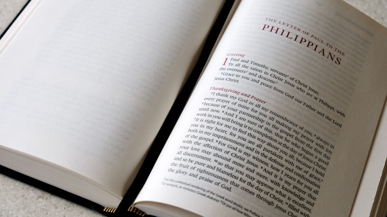An open book lying on a light-colored surface, with the left page mostly blank while the right page contains printed text. The heading 'The Letter of Paul to the Philippians' is prominently displayed at the top of the right page in capital letters. The text on the page appears to be a passage from the Bible, beginning with a large red letter 'I' for the word 'I Paul and Timothy', followed by black text with some red text headings like 'Greeting' and 'Thanksgiving and Prayer'. The book's binding is visible at the lower part of the image.