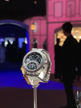 A luxurious watch with a detailed design, featuring the brand name Van Cleef & Arpels on its face. It is displayed prominently on a stand against an elegant, ambient setting with soft pink and blue lighting. Silhouettes of people are visible in the background, creating a sophisticated atmosphere.