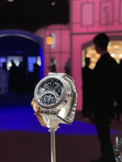A luxurious watch with a detailed design, featuring the brand name Van Cleef & Arpels on its face. It is displayed prominently on a stand against an elegant, ambient setting with soft pink and blue lighting. Silhouettes of people are visible in the background, creating a sophisticated atmosphere.
