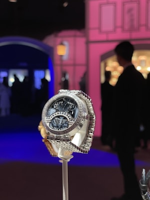 A luxurious watch with a detailed design, featuring the brand name Van Cleef & Arpels on its face. It is displayed prominently on a stand against an elegant, ambient setting with soft pink and blue lighting. Silhouettes of people are visible in the background, creating a sophisticated atmosphere.