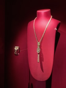 A luxurious gold necklace adorned with sparkling gems is elegantly displayed on a red velvet mannequin stand. The lighting highlights the intricate details and the lustrous finish of the jewelry, creating a sense of opulence.