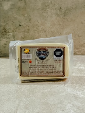 A packaged block of cheese labeled as Queso Menonita de Chihuaua with a creamy off-white color. The packaging includes detailed nutritional information and a small emblem displaying the brand logo at the top. The background is a lightly textured stone or concrete surface.