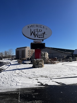 A large sign with the text 'America's Wild West' stands prominently against a clear blue sky. The sign is mounted on a stone base with some snow visible on the ground. A building is in the background, and the environment appears to be a open outdoor area with clear weather.