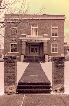 An old brick courthouse building with a symmetrical facade, featuring tall windows and a central entrance marked by a sign. The exterior landscape includes a path bordered by brick pillars leading up to several steps, and bare tree branches are visible in the foreground.