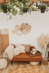 A cozy, bohemian-style seating area with a brown leather sofa adorned with various textured pillows and a knitted throw. Above the sofa, a wall features decorative woven baskets in neutral tones. The floor is wooden, complemented by round jute rugs. A wooden tray with a watering can and vase of flowers sits on the sofa. The setting is surrounded by lush greenery and pampas grass, adding a touch of nature indoors.