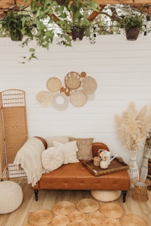 A cozy, bohemian-style seating area with a brown leather sofa adorned with various textured pillows and a knitted throw. Above the sofa, a wall features decorative woven baskets in neutral tones. The floor is wooden, complemented by round jute rugs. A wooden tray with a watering can and vase of flowers sits on the sofa. The setting is surrounded by lush greenery and pampas grass, adding a touch of nature indoors.