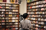 A person with short hair, wearing a striped shirt, stands in front of shelves filled with various CD covers in a store or exhibition space. The walls feature artistic posters and decorations, contributing to an eclectic and artistic atmosphere.