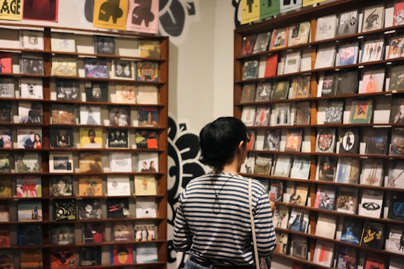 A person with short hair, wearing a striped shirt, stands in front of shelves filled with various CD covers in a store or exhibition space. The walls feature artistic posters and decorations, contributing to an eclectic and artistic atmosphere.