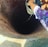 A person is lowering a bucket attached to a rope into a deep, cylindrical brick well. Water is dripping from the bucket into the well. The person's hand and part of their colorful clothing are visible. The well is built with reddish bricks.