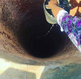 A person is lowering a bucket attached to a rope into a deep, cylindrical brick well. Water is dripping from the bucket into the well. The person's hand and part of their colorful clothing are visible. The well is built with reddish bricks.