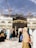 A large crowd of people, dressed mostly in traditional clothing, is walking around the Kaaba. The black cubic structure, adorned with a gold band near the top, is in the center of a spacious courtyard surrounded by a grand structure with arches and bright lights. Several cranes are visible in the background, suggesting ongoing construction amid a sky with scattered clouds.