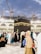 A large crowd of people, dressed mostly in traditional clothing, is walking around the Kaaba. The black cubic structure, adorned with a gold band near the top, is in the center of a spacious courtyard surrounded by a grand structure with arches and bright lights. Several cranes are visible in the background, suggesting ongoing construction amid a sky with scattered clouds.