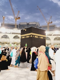 A large crowd of people, dressed mostly in traditional clothing, is walking around the Kaaba. The black cubic structure, adorned with a gold band near the top, is in the center of a spacious courtyard surrounded by a grand structure with arches and bright lights. Several cranes are visible in the background, suggesting ongoing construction amid a sky with scattered clouds.