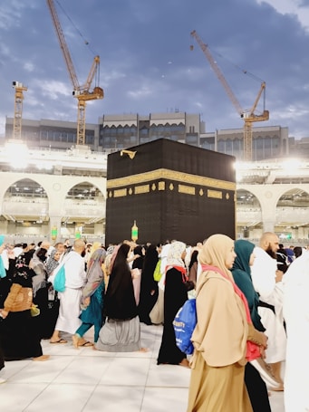 A large crowd of people, dressed mostly in traditional clothing, is walking around the Kaaba. The black cubic structure, adorned with a gold band near the top, is in the center of a spacious courtyard surrounded by a grand structure with arches and bright lights. Several cranes are visible in the background, suggesting ongoing construction amid a sky with scattered clouds.