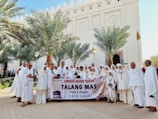 A group of people dressed in white religious attire are standing outdoors in front of a building with decorative architectural features. They are holding a banner that reads 'Umroh Akhir Tahun Talang Mas Tour & Travel' with dates '27 Des 2022 - 04 Jan 2023'. Palm trees flank the group, and everyone appears to be smiling and engaging with the camera.