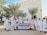 A group of people dressed in white religious attire are standing outdoors in front of a building with decorative architectural features. They are holding a banner that reads 'Umroh Akhir Tahun Talang Mas Tour & Travel' with dates '27 Des 2022 - 04 Jan 2023'. Palm trees flank the group, and everyone appears to be smiling and engaging with the camera.